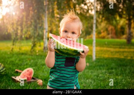 Süßer kleiner Junge, der große Slice-Wassermelone im Garten isst. Blondes Kind, das bei einem Picknick im Sommer bei Sonnenuntergang Wassermelonen im Freien isst. Gesunde Früchte Stockfoto