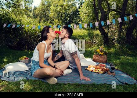Junges Paar bei einem Picknick in einem Stadtpark, das auf einer Decke sitzt, die rote Nasen trägt und sich gegenseitig küsst Selective Focus Stockfoto