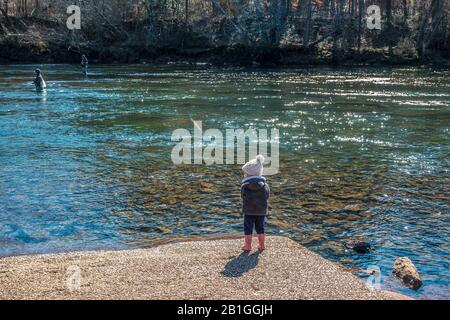 Ein kleines Mädchen, das lernt, mit einem Angelpfosten zu fischen, das geduldig wartet und an einem sonnigen Tag im Winter mit Rothe ihren rosafarbenen Hut und Gummistiefel trägt Stockfoto