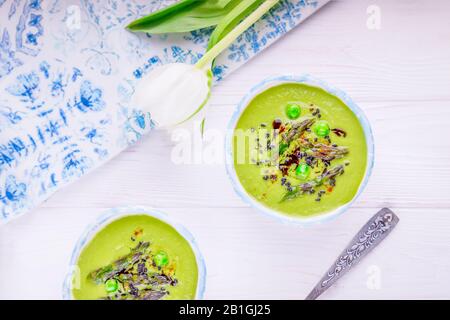 Spargelsaison. Hausgemachte grüne Sahnesuppe mit Spargel, grünen Erbsen, schwarzen Sesamsamen und Kürbisöl auf weißem Grund Stockfoto
