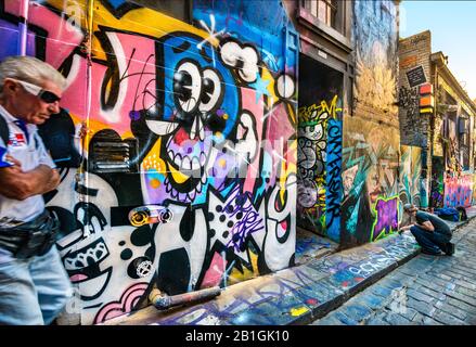 Mann mit Sonnenbrille, die an der Seite des Kopfsteinpflasterwegs steht, während junger Mann Graffiti, Hosier Street, Melbourne Lanes, Melbourne, Vic malt Stockfoto