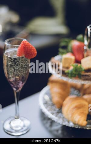 Eine Nahaufnahme einer Erdbeere auf einem Champagnerglas mit Croissants Erdbeeren und Kuchen auf einem gestuften Tortenständer im Hintergrund Stockfoto