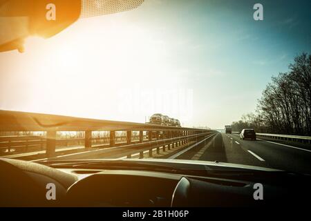 Autofensteransicht von LKWs, die auf der Autobahn mit hinterem Sonnenschein fahren - schnelle Fahrzeuge bei Sonnenuntergang - Transportkonzept - Schwerpunkt auf Halbzeug und Stockfoto