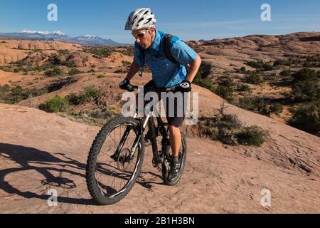 25. Februar 2020: Das Bureau of Land Management kündigte am 24. Februar an, zwei Parzellen innerhalb des Sand Flats Special Recreation Area in der Nähe von Moab, Utah, aus einem bevorstehenden Verkauf von Öl- und Gaspacht zu entfernen. Der Plan hätte Sondierungsbohrungen unter dem Slickrock Trail ermöglicht, einem der beliebtesten Mountainbike-Ziele der Welt. Bild: 5. April 2015 - Moab, Utah, USA - Mountain Biker TIM-SPUR, die den roten Sandstein des berühmten Slickrock-Trails mit La Sal Bergen und Schnee hinter sich führt. Moab bietet einige der spektakulärsten und anspruchsvollsten Mountainbike-Touren an. Po Stockfoto