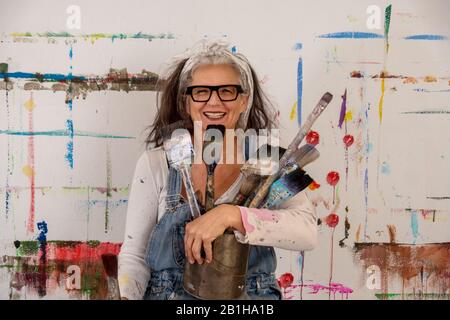 Lächelnde ältere Frau, stolze Künstlerin, in ihren fünfziger Jahren mit grauem Haar und schwarzer Brille und vielen Pinsel Stockfoto