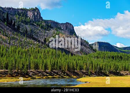 Fluss, der durch grüne Wiesen mit grünem Wald fließt, der unter blauem Himmel und weißen Wolken dem felsigen Berghang Platz gibt. Hier ist keine Maske erforderlich. Stockfoto