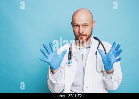 Attraktiver Mann kahler bärtiger Arzt in medizinischen Gummihandschuhen, der die Kamera auf dem blauen Hintergrund isoliert betrachtet Stockfoto