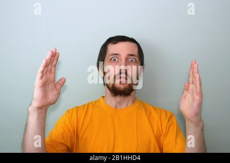 Ein Mann mittleren Alters mit einem Schnurrbart und einem brünetten Bart in einem gelben T-Shirt auf grauem Hintergrund zeigt etwas großes zwischen den Handflächen, das seine Hände vor ihm spreizt. Stockfoto