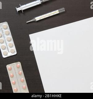 Vorlage für weißes Papier mit Pillen in Blister, Thermometer und Spritze zur Injektion auf dunklem, wengem Holzhintergrund. Die Bereitstellung medizinischer und pharmakologischer Dienstleistungen. Stockfoto