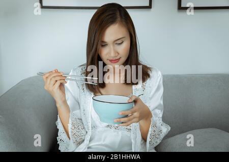 Thailändische Frauen essen nachts Essen. Asiatische Mädchen in weißer Nachtgown und langärmeliger Satin-Robe mit Blumenspitze können wegen Hunger im Leben nicht schlafen Stockfoto