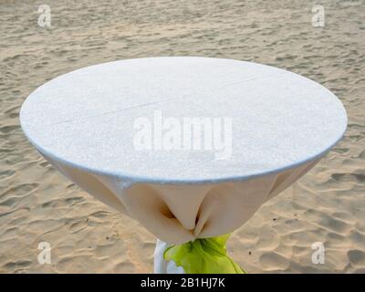 Leerer runder Esstisch mit weißer Tischdecke und hellgrüner Schleife am Strand. Stockfoto