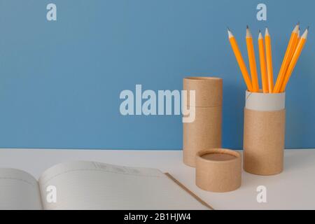 Papierröhrchen mit gelben Bleistiften und Notizbuch auf weißem Tisch auf blauem Hintergrund. Recycling-Konzept, das zeigt, wie Papierrohre zu Hause oder im Büro wiederverwendet werden Stockfoto