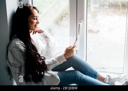 Attraktive junge lächelnde Frau, die mit einem Handy auf der Fensterbank sitzt