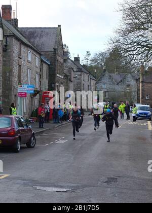 Frauen Rennen auf der Hauptstraße des Dorfes mit Pfannen und Pfannkuchen während Winster Pancake Races Shrove Tuesday Pancake Day Derbyshire UK Stockfoto