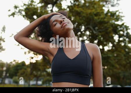 Porträt einer aktiven afroamerikanischen jungen Frau, die an einem sonnigen Tag schwarzen Sport-BH mit geschlossenen Augen trägt und den Hals im Park streckt Stockfoto