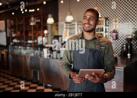 Fröhlicher junger männlicher Besitzer, der digitales Tablet hält, während er im Café steht Stockfoto