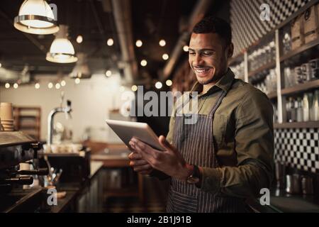 Portrait des erfolgreichen jungen afro-amerikanischen Café-Besitzers, der hinter der Theke steht und digitale Tabletts verwendet Stockfoto