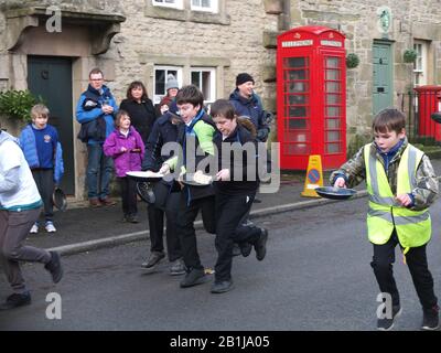 Schulkinder Rennen auf der Hauptstraße des Dorfes mit Pfannen und Pfannkuchen während Winster Pancake-Rennen am Dienstag Shrove Pancake Day Derbyshire UK Stockfoto