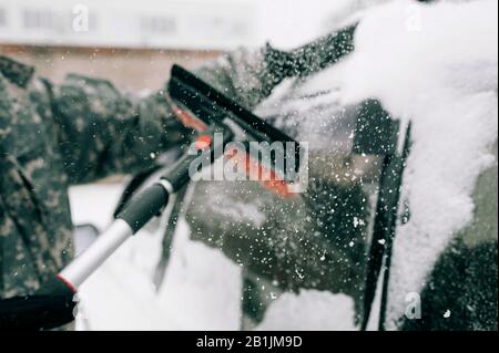 Der Mann in Tarnkleidung ohne Gesicht reinigt das Auto mit einem schwarzen Pinsel mit rotem Haufen aus schwerem Schnee. Reinigung des schneebedeckten Glases der Ca Stockfoto