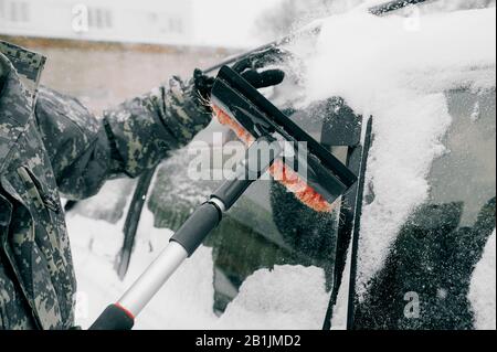 Der Mann in Tarnkleidung ohne Gesicht reinigt das Auto mit einem schwarzen Pinsel mit rotem Haufen aus schwerem Schnee. Reinigung des schneebedeckten Glases der Ca Stockfoto