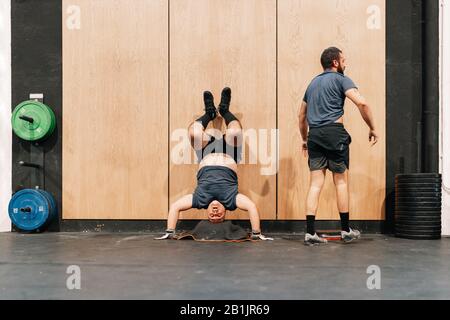 Zwei Männer trainieren auf dem Boden in einem Fitnessstudio, das eine Wand mit Handstand macht Stockfoto