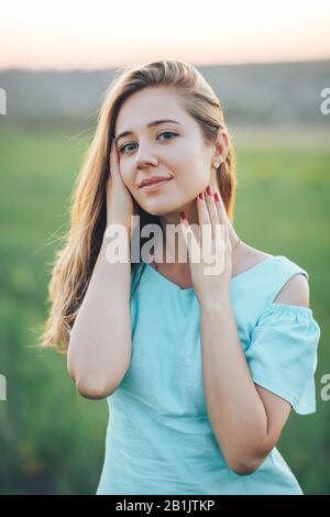 Schönes Mädchen in einem blauen Kleid mit langen, gewellten Haaren und bezaubernden Augen, die auf die Kamera blicken und ihr Gesicht mit beiden Händen berühren Stockfoto