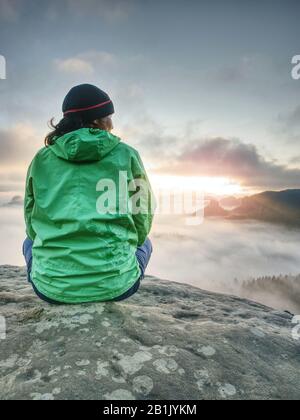 Grüne Jacke Frau sitzt penibel am Rand eines Felsens und betrachtet die nebligen Wolken. Stockfoto