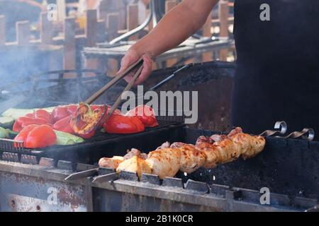 Küchenchef brat Fleisch und Gemüse auf dem Grill. In schwarzer Schürze geröstete rote Paprika und Zucchini kochen. Grill im Freien. Stockfoto