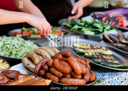 Grillwürstchen auf der Straße. Straßenküche und Kochkonzept für den Außenbereich. Hände von Frauen mit Tangas im Hintergrund. Stockfoto