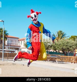 Ein gruseliger Clown, der ein farbenfrohes gelbes, rotes und blaues Kostüm trägt und auf einem öffentlichen Spielplatz im Freien springt Stockfoto