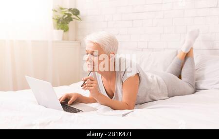 Pensive Seniorin beim Surfen auf einem Laptop im Bett Stockfoto