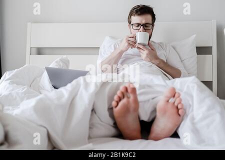 Entspannter Mann, im Schlafanzug, trinkt aromatischen Kaffee, sitzt im Bett mit den Füßen aus dem Boden, ein Laptop liegt in der Nähe. Stockfoto