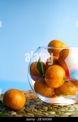 Orangefarbene reife Mandarinen mit grünen Blättern in einer runden Glasschale auf hellblauem Grund. Vertikales Format. Stockfoto