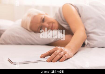 Müde ältere Frau im Bett, die Zeit auf ihrem Telefon überprüft Stockfoto