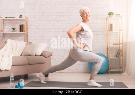 Sportliche Seniorin macht Ausfallschritte zu Hause, leer Stockfoto
