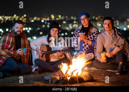 Eine Gruppe von Leuten, die nachts in der Nähe von Lagerfeuer sitzen und Gitarre spielen, Lieder singen und glücklich miteinander sprechen. Stockfoto