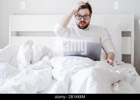 Der Mann mit einem Laptop im Bett erhielt schlechte Nachrichten, er ist verwirrt, hält die Hand am Kopf und beobachtet den Bildschirm in schockierten Augen. Stockfoto