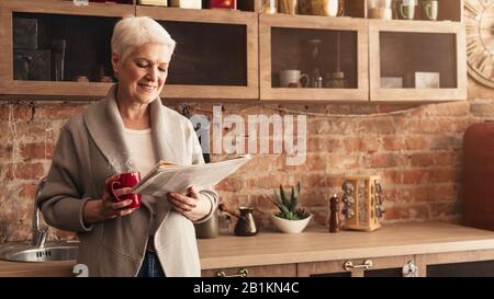 Fröhliche Ältere Frau Liest Zeitung Und Trinkt Heißen Tee In Der Küche Stockfoto