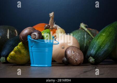 Gemüse. Gemüse liegt auf einem Holztisch, darunter Kürbisse, Kürbisse, Zucchini und Pilze. Im Vordergrund steht ein kleiner blauer Eimer Stockfoto