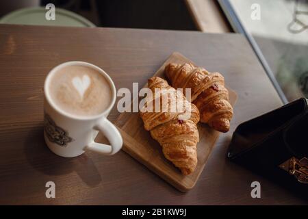 Cappuccino mit schöner Latte und Croissant auf Holzhintergrund auf dem Tisch. Perfektes Frühstück am Morgen. Rustikaler Stil. Stockfoto