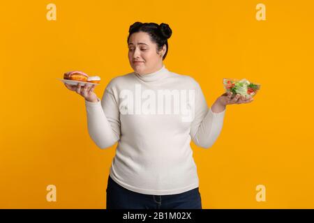 Unsicheres Plump-Mädchen kann sich nicht zwischen Donuts und frischem Gemüsesalat entscheiden Stockfoto