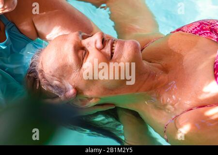 Fröhliche lächelnde Seniorin, die mit einer Kopfmassage im Wasserbad mit einem kochenden Lächeln und geschlossenen Augen im Swimmingpool eine entspannende, frische Atmosphäre erhält Stockfoto