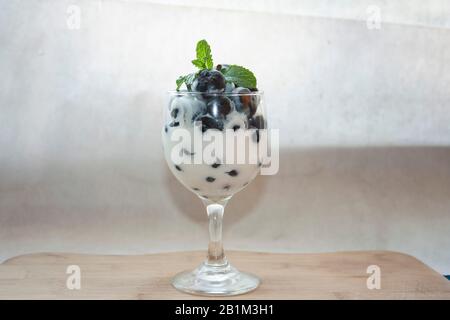 Tasse mit kaltem, naturweißem Joghurt, gemischt mit Blaubeeren und frischen Zierblättern aus Minzenblatt auf Holzbrett mit Trio Jumbo Fruits und Star at Stockfoto