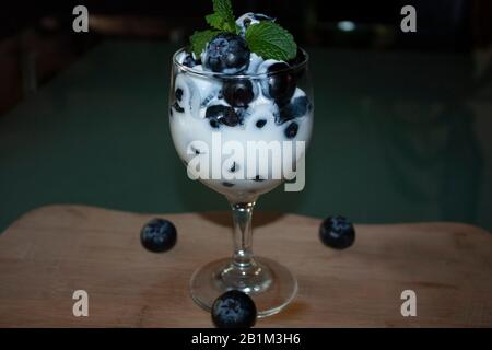 Tasse mit kaltem, weißem Joghurt, gemischt mit Blaubeeren und frischen Zierblättern aus Minzenblatt auf Holzbrett, umgeben von drei Blaubeeren Stockfoto