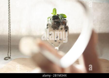 Tasse mit kaltem, naturweißem Joghurt, gemischt mit Blaubeeren und frischen Zierblättern aus Minzenblatt, auf Holztisch mit hellem Herzen gerahmt Stockfoto
