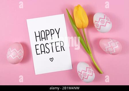"Happy Easter"-Textkarte umgeben von ostereiern und einzelnen gelben Tulpenfrüchteblumen auf rosa gefärbtem Hintergrund, flach gelegt Stockfoto