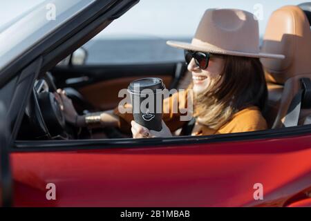Junge stilvolle Frau im Hut fahren Cabrio Auto in der Nähe des Ozeans, genießen Sommerferien, Kaffee trinken. Konzept einer unbeschwerten Reise und Sommerzeit Stockfoto