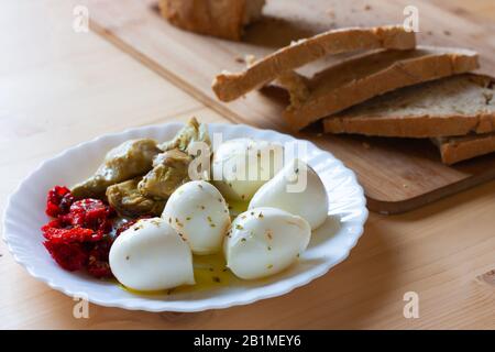 Mozzarella, Kirschtomaten getrocknet und Artischocken gewürzt mit Olivenöl und Oregano auf weißem Teller mit Brotscheiben auf Holzschneidebrett Stockfoto