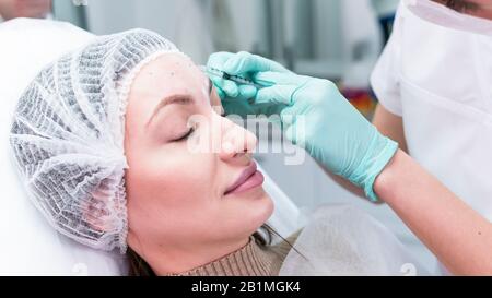 Die junge Kosmetikerin, die sich auf die Injektion in die weibliche Stirn vorbereitet. Der Arzt Kosmetologe macht Anti-Aging-Behandlung und Gesichtsaufhebung probed Stockfoto