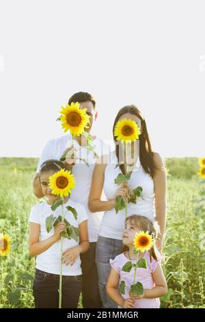 Glückliche Familie von Eltern und zwei Mädchen, die sich umarmen, während sie Sonnenblumen halten und im Sommerfeld mit der Kamera lächeln Stockfoto